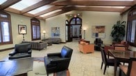 This distinguished single-floor space presents a sophisticated lounge or lobby environment defined by strong architectural character and thoughtful interior design. The most striking overhead feature is the vaulted ceiling accented by rich, dark wood beams that run the length of the room, lending warmth and a sense of grandeur to the open layout. Below, travertine tile flooring in neutral tones anchors the space with an air of timeless elegance.

A grand arched entryway with dark wood detailing serves as a dramatic focal point, framing the threshold and drawing the eye toward additional interior space beyond. Multiple seating arrangements are distributed throughout, including plush leather armchairs, striped upholstered sofas, and contemporary accent chairs in warm orange tones, creating distinct conversation areas suited for both relaxed lounging and casual meetings.

Natural light filters in through plantation-shuttered windows, complemented by floor lamps that cast a warm, ambient glow throughout the room. Curated abstract artwork adorns the walls, adding a modern, creative touch to the otherwise classic architectural backdrop. The overall ambiance balances formal design elements with inviting comfort, making this a versatile and visually compelling gathering space.