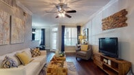 This single-floor residence offers a thoughtfully designed layout with distinct living, dining, kitchen, bedroom, and bathroom areas. The living room serves as the heart of the home, showcasing rich hardwood flooring, crown molding, decorative columns, and a ceiling fan. Abundant natural light filters through large windows dressed with navy drape panels and white blinds, creating a bright and inviting atmosphere. The kitchen is outfitted with dark cabinetry, granite countertops, hardwood flooring, and stainless steel appliances, providing both functionality and a sleek aesthetic. Adjacent to the living area, the dining space accommodates a round wood table set beneath a statement pendant light, with a large window enhancing the airy feel. The bedroom features plush carpeting, crown molding, a ceiling fan, and a generous window that allows natural light to fill the room. A dedicated sitting area with carpet connects directly to the bathroom, which includes a full soaking tub with white tile surround. Throughout the residence, a cohesive neutral color palette with warm gold accents and carefully chosen artwork contributes to a polished and comfortable living environment.