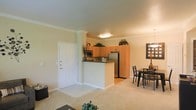 This single-floor residence is designed with an open-concept layout that seamlessly connects the kitchen, dining area, and living space. The kitchen is positioned centrally and features warm wood-toned cabinetry that extends to the ceiling, providing ample storage. A peninsula with a dark countertop serves as a natural divider between the kitchen and the surrounding living areas, offering additional workspace and a casual gathering point. Stainless steel appliances, including a refrigerator and microwave, add a modern touch to the functional kitchen setup. The flooring transitions from tile in the kitchen and entryway to plush carpet in the living and dining areas, defining each zone while maintaining visual continuity. Crown molding runs along the ceiling throughout, lending an elevated, polished character to the interior. Neutral wall tones keep the space feeling bright and versatile. The dining area accommodates a full table and chair set, positioned beneath a decorative pendant light. The front entry door is prominently placed and finished in white with classic paneling. Overall, the space presents a practical yet refined layout suited to everyday living.
