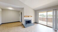 This single-story unit presents a well-organized layout with a thoughtful mix of finishes throughout. The living room serves as the focal point of the space, showcasing vaulted ceilings that lend an open, airy quality to the room. A wood-manteled fireplace with a gray brick hearth anchors one wall, while a large sliding glass door opposite floods the area with natural light and provides direct access to a private balcony with outdoor views. Light wood-style flooring runs throughout the living area and entryway, complementing the neutral gray walls and dark baseboards.

The kitchen is outfitted with slate-blue cabinetry, light quartz-style countertops, a stainless steel refrigerator, and a full-length windowed door that brings in additional natural light. The bedrooms feature plush wall-to-wall carpeting and ceiling fans, offering a comfortable and functional retreat. One bedroom includes a generously sized walk-in closet with built-in shelving and hanging rods for ample storage. Throughout the unit, consistent dark-trimmed doorways and a neutral color palette create a cohesive, modern aesthetic.