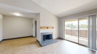 This single-story unit presents a well-organized layout with a thoughtful mix of finishes throughout. The living room serves as the focal point of the space, showcasing vaulted ceilings that lend an open, airy quality to the room. A wood-manteled fireplace with a gray brick hearth anchors one wall, while a large sliding glass door opposite floods the area with natural light and provides direct access to a private balcony with outdoor views. Light wood-style flooring runs throughout the living area and entryway, complementing the neutral gray walls and dark baseboards.

The kitchen is outfitted with slate-blue cabinetry, light quartz-style countertops, a stainless steel refrigerator, and a full-length windowed door that brings in additional natural light. The bedrooms feature plush wall-to-wall carpeting and ceiling fans, offering a comfortable and functional retreat. One bedroom includes a generously sized walk-in closet with built-in shelving and hanging rods for ample storage. Throughout the unit, consistent dark-trimmed doorways and a neutral color palette create a cohesive, modern aesthetic.