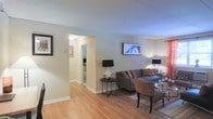 This single-floor residence offers a clean, functional layout designed for comfortable everyday living. The living area serves as the heart of the home, showcasing warm light hardwood flooring that extends throughout the space, creating a cohesive and inviting atmosphere. Neutral wall tones in soft grey and white provide a versatile backdrop that enhances the sense of openness and brightness throughout.

The living room is thoughtfully arranged with a plush grey sofa, coordinating accent chairs, and stylish side tables, all anchored by a patterned area rug. A wall-mounted flat-screen TV adds a modern touch, while carefully placed floor and table lamps provide warm ambient lighting that complements the natural light filtering into the room.

Crown moulding runs along the ceiling, adding a refined architectural detail that elevates the overall finish of the space. A wide doorway connects the living area to the bedroom, which is visible in the background and features similar flooring and a mirrored wardrobe, suggesting a consistent and well-considered design throughout. Clean white baseboards and trim frame each room neatly, contributing to the polished, move-in-ready character of this welcoming residence.