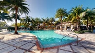 This inviting outdoor amenity space centers around a generously sized swimming pool with pristine, crystal-clear turquoise water. The pool features a gently sloping entry with handrails, making it accessible and welcoming. The surrounding deck is finished with a combination of decorative brick-edged pavers and stone tile, creating an attractive and durable surface for lounging and entertaining.

A covered cabana or pergola structure anchors one side of the pool area, constructed in a Mediterranean-inspired architectural style with stucco walls and a tiled roof. The structure provides shaded seating with lounge chairs positioned to overlook the pool. Additional seating areas with umbrellas are visible throughout the deck, offering ample space for relaxation.

The entire area is enveloped by a lush tropical landscape, with tall, mature palm trees framing the space and creating a resort-like ambiance. Lush greenery and tropical plantings enhance the sense of seclusion and leisure. The open sky above allows for abundant natural sunlight, complementing the vibrant outdoor atmosphere. In the background, residential buildings are visible, nestled within the verdant surroundings, reinforcing the community-oriented nature of this well-appointed outdoor amenity.