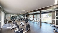 Located at 1602 Green Mountain Dr in Little Rock, Arkansas, this fitness center offers a well-equipped, single-floor exercise facility designed to support a comprehensive workout experience. The space is outfitted with a diverse selection of cardio machines, including upright and recumbent stationary bikes, ellipticals, stair steppers, and treadmills, as well as strength training equipment. A full-length mirrored wall runs along one side of the room, visually expanding the space and allowing users to monitor their form during workouts. The flooring throughout is durable and exercise-appropriate, providing a stable surface for a variety of fitness activities. Large sliding windows and French doors line the exterior walls, flooding the space with natural light and offering views of the surrounding outdoor area, including what appears to be a tennis or sports court. The walls feature a dark wood panel design with white trim accents, giving the facility a clean, structured aesthetic. Overhead fluorescent strip lighting ensures consistent illumination throughout. A bulletin board near the entrance provides an informational focal point for users. The layout is open and organized, allowing for comfortable movement between equipment stations.