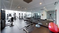This fitness center is a comprehensively outfitted facility designed to support a full range of workout routines. The space is defined by its clean, modern aesthetic, featuring a dropped tile ceiling with recessed lighting, neutral white walls, and durable rubber flooring throughout.

A standout architectural feature is the expansive floor-to-ceiling glass windows along one wall, which flood the space with natural light and offer sweeping views of the surrounding urban skyline — creating an energizing atmosphere for workouts.

Large wall-to-wall mirrors are strategically placed throughout the gym, visually expanding the space and allowing users to monitor their form. The cardio zone is well-stocked with a row of treadmills, elliptical trainers, and a recumbent bike, many equipped with individual digital displays.

The free weights area includes a fully stocked dumbbell rack alongside flat and adjustable benches, catering to a variety of strength training exercises. Complementing this is a multi-station cable and weight machine, ideal for targeted resistance training. Additional functional training tools, such as BOSU balance trainers and exercise mats, are also present.

The layout is thoughtfully organized, with distinct zones for cardio, free weights, and strength machines, ensuring efficient use of the space. An access door provides a private entry point, reinforcing the facility's role as a dedicated, professional-grade fitness environment.