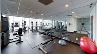 This fitness center is a comprehensively outfitted facility designed to support a full range of workout routines. The space is defined by its clean, modern aesthetic, featuring a dropped tile ceiling with recessed lighting, neutral white walls, and durable rubber flooring throughout.

A standout architectural feature is the expansive floor-to-ceiling glass windows along one wall, which flood the space with natural light and offer sweeping views of the surrounding urban skyline — creating an energizing atmosphere for workouts.

Large wall-to-wall mirrors are strategically placed throughout the gym, visually expanding the space and allowing users to monitor their form. The cardio zone is well-stocked with a row of treadmills, elliptical trainers, and a recumbent bike, many equipped with individual digital displays.

The free weights area includes a fully stocked dumbbell rack alongside flat and adjustable benches, catering to a variety of strength training exercises. Complementing this is a multi-station cable and weight machine, ideal for targeted resistance training. Additional functional training tools, such as BOSU balance trainers and exercise mats, are also present.

The layout is thoughtfully organized, with distinct zones for cardio, free weights, and strength machines, ensuring efficient use of the space. An access door provides a private entry point, reinforcing the facility's role as a dedicated, professional-grade fitness environment.