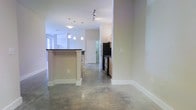 This single-floor space showcases a clean, modern aesthetic defined by polished concrete floors that run throughout the open-concept layout, lending the interior an industrial yet refined character. The kitchen serves as a central feature, equipped with stainless steel appliances including a refrigerator and range, warm wood-toned cabinetry, and a peninsula island topped with a dark countertop — ideal for both meal preparation and casual dining. A row of pendant lights hangs above the island, complementing the track lighting overhead to create a layered and well-lit environment. The open flow between the kitchen and adjacent living areas allows for a spacious, unobstructed feel. White walls and bright white trim provide a crisp contrast to the warm tones of the cabinetry and the cool gray of the concrete underfoot. A white paneled door leads toward additional areas of the unit, maintaining a cohesive and tidy design throughout. Natural light filters in through windows at the far end of the space, further enhancing the bright and airy ambiance. Overall, the space combines functional design with contemporary finishes for a polished, livable environment.