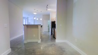 This single-floor space showcases a clean, modern aesthetic defined by polished concrete floors that run throughout the open-concept layout, lending the interior an industrial yet refined character. The kitchen serves as a central feature, equipped with stainless steel appliances including a refrigerator and range, warm wood-toned cabinetry, and a peninsula island topped with a dark countertop — ideal for both meal preparation and casual dining. A row of pendant lights hangs above the island, complementing the track lighting overhead to create a layered and well-lit environment. The open flow between the kitchen and adjacent living areas allows for a spacious, unobstructed feel. White walls and bright white trim provide a crisp contrast to the warm tones of the cabinetry and the cool gray of the concrete underfoot. A white paneled door leads toward additional areas of the unit, maintaining a cohesive and tidy design throughout. Natural light filters in through windows at the far end of the space, further enhancing the bright and airy ambiance. Overall, the space combines functional design with contemporary finishes for a polished, livable environment.