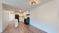 This single-floor unit offers a clean, modern aesthetic throughout. The kitchen serves as a functional centerpiece, featuring crisp white shaker-style cabinetry extending to the ceiling for ample storage, light countertops, a stainless single-basin sink with a matte black faucet, and a suite of black appliances including a gas range and refrigerator. A kitchen island adds additional prep space and a natural transition into the adjacent living area. The living room is bright and open, with warm wood-look flooring that flows from the kitchen and transitions to plush neutral carpet, creating a comfortable and defined space. Vertical blinds on the main window allow for adjustable natural light. A hallway connects the living areas to the private spaces, where the carpeted bedrooms offer a quiet retreat. Each bedroom features neutral tones, window access to natural light, and ample floor space. The bathroom is thoughtfully updated with a white vanity, light countertop, a large mirror, wood-look flooring, and clean white tile wainscoting. The overall color palette of soft grays, whites, and warm wood tones gives the unit a cohesive and contemporary feel throughout.