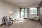 This single-floor unit is laid out to maximize functional living space across several distinct areas. The living room is generously sized, featuring neutral-toned carpet, a textured popcorn ceiling, dark wood baseboards, and a ceiling fan. Large windows allow natural light to filter in, offering views of surrounding greenery, while a wall-unit HVAC system provides climate control. The entry area transitions between hard flooring and carpet, framed by a solid wood front door with wall sconce lighting nearby.

The galley-style kitchen is equipped with rich, dark wood cabinetry extending to the ceiling, a double-basin stainless steel sink, light-toned countertops, a built-in microwave, dishwasher, and a range. Hard wood-look flooring runs throughout the kitchen, providing a practical and clean surface.

Both bedrooms are carpeted and well-lit by windows with outdoor views, each featuring a wall-unit HVAC. The layout, visible in the floor plan overview, shows a corridor connecting the bedrooms to two bathrooms. Each bathroom includes a wood-tone vanity with a white countertop, a mirrored medicine cabinet, and hard flooring, with one bathroom featuring a shower-tub combination.