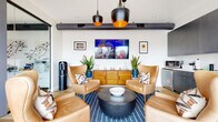 This thoughtfully designed single-floor space blends modern industrial aesthetics with warm, inviting furnishings. The centerpiece of the lounge area features a curated arrangement of tan leather wingback chairs surrounding a dark hammered-metal coffee table, all anchored by a bold navy-striped area rug. Striking matte-black pendant lights with copper interiors hang from an exposed ductwork ceiling, casting a warm glow over the seating area. A wall-mounted flat-screen television is flanked by lush potted plants and framed artwork, with a mid-century style wooden credenza beneath adding a natural, earthy tone. Light hardwood flooring runs throughout, enhancing the bright, airy atmosphere created by white walls and recessed lighting. A sleek glass partition wall connects the lounge to an adjacent room, maintaining an open and connected feel. The kitchenette features rich dark wood cabinetry, light stone countertops, a stainless steel sink, microwave, and under-counter beverage refrigerator — ideal for hosting. A clean, accessible restroom with large-format floor tiles, crisp white subway wall tiles, matte black hardware, and a modern vanity with gold accents completes the space. Every detail reflects a cohesive, contemporary design sensibility.