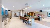 Located on the third floor at 3099 Iowa Ave in Riverside, California, this 2,180-square-foot resident lounge is a thoughtfully designed amenity space offering a variety of distinct functional zones within an open, modern layout. Large-format light tile flooring spans the main areas, complemented by warm wood-toned accents throughout. Vertical slatted wood partitions elegantly divide the space into semi-private zones without fully enclosing them, maintaining an open and airy atmosphere. A central dining area features a round wood table with blue accent chairs beneath a dome pendant light, ideal for casual gatherings. Adjacent to this is a well-appointed wet bar and kitchenette with a raised island, bar-height seating, and dual wall-mounted flat-screen TVs backed by wood-panel and tile finishes. Stylish flush-mount and pendant lighting with gold-interior shades add warmth throughout. A relaxed lounge area provides comfortable seating, while a dedicated gaming station with a lit arcade-style table and bar stools offers entertainment. Floor-to-ceiling windows with roller shades and multiple glass doors flood the space with natural light and provide direct access to an outdoor terrace. The overall ambiance is contemporary and upscale, designed to foster community and comfort for residents.