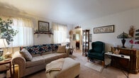 This single-floor residence offers a practical and efficiently organized layout, encompassing a living room, a combined dining room and kitchen, one bedroom, one bathroom, and a hallway. The carpeted living room serves as the central gathering space, featuring large windows dressed with sheer curtains that allow natural light to fill the room. A decorative wall shelf adds character, while the open sightline toward the bedroom hallway enhances the sense of flow throughout the space. The entry hallway is welcoming, with vaulted accent walls and a gallery of framed photographs that give it a personalized, homey feel. The combined kitchen and dining area is both functional and efficient. The kitchen features warm honey-toned wood cabinetry, ample upper and lower storage, a full range, refrigerator, and a countertop workspace. The adjacent dining area accommodates a full dining table and chairs, with decorative shelving adding a charming, cottage-like aesthetic. The bedroom is a comfortable retreat with carpeted floors, a window allowing natural light, and sufficient space for a full bed and additional storage furnishings. The bathroom is clean and well-maintained, featuring a shower-tub combination with a bold patterned curtain, a wood-toned vanity with countertop sink, and a medicine cabinet with mirror.