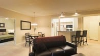 This single-floor residence offers an open-concept layout that seamlessly connects the living, dining, and kitchen areas into one cohesive space. The living area is anchored by a dark leather sofa paired with rich burgundy accent pillows, complemented by light-toned flooring and warm beige walls that create an inviting atmosphere. A large framed mirror on the living room wall adds depth and enhances the sense of space.

Adjacent to the living area, the dining space features a round wooden table with matching chairs, illuminated by an elegant chandelier that provides warm, focused lighting. This area benefits from its open connection to the rest of the space, making it ideal for everyday dining or entertaining.

The kitchen is accessible through a defined passthrough opening, featuring a raised breakfast bar with bar stool seating, dark countertops, and white cabinetry. Modern appliances, including a microwave and range, are efficiently integrated into the layout. Recessed and track lighting throughout the kitchen ensures a well-lit workspace. Overall, the space maintains a cohesive, warm aesthetic with functional design elements.