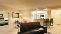 This single-floor residence offers an open-concept layout that seamlessly connects the living, dining, and kitchen areas into one cohesive space. The living area is anchored by a dark leather sofa paired with rich burgundy accent pillows, complemented by light-toned flooring and warm beige walls that create an inviting atmosphere. A large framed mirror on the living room wall adds depth and enhances the sense of space.

Adjacent to the living area, the dining space features a round wooden table with matching chairs, illuminated by an elegant chandelier that provides warm, focused lighting. This area benefits from its open connection to the rest of the space, making it ideal for everyday dining or entertaining.

The kitchen is accessible through a defined passthrough opening, featuring a raised breakfast bar with bar stool seating, dark countertops, and white cabinetry. Modern appliances, including a microwave and range, are efficiently integrated into the layout. Recessed and track lighting throughout the kitchen ensures a well-lit workspace. Overall, the space maintains a cohesive, warm aesthetic with functional design elements.