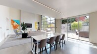 This contemporary single-floor space showcases a striking open-plan design defined by soaring ceilings, polished concrete floors, and an abundance of natural light. The living area serves as the heart of the space, anchored by a sleek wall-mounted fireplace topped with a flat-screen TV and flanked by bold, large-format pop-art canvases that add vivid color and personality. A full-height industrial-style glass garage door opens the living zone directly to a private outdoor patio, seamlessly blending interior and exterior living. A sliding glass door provides an additional connection to the landscaped outdoor area.

The dining area features a large white rectangular table surrounded by upholstered chairs, positioned beneath a linear recessed ceiling light that runs the length of the room. A secondary lounge area offers a pair of wingback chairs accented with chartreuse cushions, set against a wall of sleek white built-in cabinetry with under-cabinet lighting.

The chef's kitchen is a standout feature, equipped with white flat-front cabinetry, a vibrant yellow subway tile backsplash, a large central island with bar seating, a gas cooktop, double wall ovens, and a bold matte black refrigerator. A stainless steel range hood completes the professional-grade setup. Throughout, the design maintains a cohesive, modern aesthetic with a crisp neutral palette punctuated by energetic yellow accents.