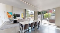 This contemporary single-floor space showcases a striking open-plan design defined by soaring ceilings, polished concrete floors, and an abundance of natural light. The living area serves as the heart of the space, anchored by a sleek wall-mounted fireplace topped with a flat-screen TV and flanked by bold, large-format pop-art canvases that add vivid color and personality. A full-height industrial-style glass garage door opens the living zone directly to a private outdoor patio, seamlessly blending interior and exterior living. A sliding glass door provides an additional connection to the landscaped outdoor area.

The dining area features a large white rectangular table surrounded by upholstered chairs, positioned beneath a linear recessed ceiling light that runs the length of the room. A secondary lounge area offers a pair of wingback chairs accented with chartreuse cushions, set against a wall of sleek white built-in cabinetry with under-cabinet lighting.

The chef's kitchen is a standout feature, equipped with white flat-front cabinetry, a vibrant yellow subway tile backsplash, a large central island with bar seating, a gas cooktop, double wall ovens, and a bold matte black refrigerator. A stainless steel range hood completes the professional-grade setup. Throughout, the design maintains a cohesive, modern aesthetic with a crisp neutral palette punctuated by energetic yellow accents.