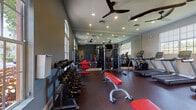 This Athletic Club fitness center is a thoughtfully designed, single-floor facility built to support a full range of workout routines. The space features durable rubber flooring throughout, providing both comfort and safety for exercise. Large arched and rectangular windows line the walls, filling the room with abundant natural light and offering views of the surrounding outdoor greenery, while ceiling fans and recessed lighting ensure a comfortable and well-lit environment at all times.

Full-length mirrors span an entire wall, visually expanding the space and allowing users to monitor their form during training. The cardio zone is equipped with multiple treadmills, an elliptical trainer, and a stair climber, catering to a variety of cardiovascular workouts. A central multi-functional cable machine serves as the focal point of the strength training area, complemented by a comprehensive dumbbell rack with an extensive range of free weights and adjustable workout benches with red upholstery. The cool gray walls and dark flooring give the facility a modern, professional aesthetic. The space is organized to allow for smooth traffic flow between equipment zones, making it practical and user-friendly for individuals at all fitness levels.