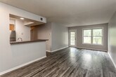 This single-floor residence offers a well-organized layout that balances open communal areas with private living spaces. The main living area is spacious and welcoming, finished with dark hardwood-style flooring that extends throughout the common areas. Large windows and a glass-paneled door flood the space with natural light while providing views of the surrounding greenery, creating a bright and airy atmosphere.

The kitchen is thoughtfully designed in a galley style, featuring warm honey-toned cabinetry, dark countertops, a double stainless steel sink, and a full suite of stainless steel appliances including a refrigerator, electric range, built-in microwave, and dishwasher. A raised breakfast bar with dark countertop serves as a functional partition between the kitchen and the main living area, enhancing the open feel while defining each zone.

The bedrooms are carpeted for comfort and each benefits from natural light provided by well-positioned windows. Closet space is included, adding practical storage. A hallway connects the sleeping areas to the bathroom, which features a warm wood-toned vanity with ample cabinet storage, a well-lit mirror, and a full bathtub with chrome fixtures. The overall palette of soft gray walls and neutral finishes gives the space a clean, consistent, and modern character throughout.