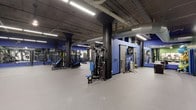 This single-floor fitness facility offers a generous open layout designed to accommodate a comprehensive workout experience. The space is defined by its industrial aesthetic, featuring an exposed ceiling with visible ductwork, conduit, and structural piping finished in dark tones, lending the gym a modern, urban character. Rows of linear fluorescent lighting illuminate the floor evenly, ensuring excellent visibility throughout.

The gym is equipped with a robust selection of strength-training machines, including cable and pulley systems with blue upholstered seating, free weight benches, and various resistance equipment positioned throughout the open floor plan. Structural columns painted in matte black are integrated into the layout, adding to the industrial design while naturally segmenting workout zones.

Bold blue accent walls provide striking visual contrast against the darker overhead elements and serve as a functional backdrop for wall-mounted mirrors, which enhance the sense of depth and allow users to monitor their form during exercise. The floor is finished with grey rubber sport flooring, offering durability, impact absorption, and safety. Overall, the space delivers a focused, professional training environment with a sleek, contemporary edge.