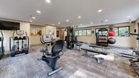 This dedicated fitness center offers a generously proportioned, single-room workout space designed to accommodate a full range of exercise needs. The room features a neutral-toned interior with warm beige walls, a smooth white ceiling lined with recessed lighting, and durable patterned carpet flooring that provides both comfort and traction underfoot. Large, black-framed windows span multiple walls, flooding the space with natural light and offering pleasant views of surrounding greenery and mature trees, creating an inviting and energizing atmosphere. The strength-training area is well-appointed with a multi-station cable machine, a set of weight benches, and a full dumbbell rack stocked with a wide range of free weights. The cardio zone features an array of equipment including treadmills, elliptical trainers, a rowing machine, a stationary cycle, and a stair climber, ensuring comprehensive cardiovascular training options. Motivational fitness artwork is displayed along the walls, adding a polished, purposeful aesthetic to the space. The layout is thoughtfully organized to allow ample circulation between equipment. A dedicated TRX suspension training station and medicine ball storage further round out the fitness offerings, making this a fully functional and versatile facility.