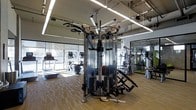 This contemporary fitness center is thoughtfully designed to support a full range of training activities. The space is anchored by a professional-grade multi-functional cable machine at its center, offering a versatile platform for strength and resistance training. Surrounding it, an array of additional strength equipment and cardio machines — including treadmills and ellipticals — are arranged to maximize usability and flow throughout the floor.

The interior benefits from abundant natural light streaming through expansive floor-to-ceiling windows along the perimeter, creating an open and energizing atmosphere. The light wood-toned flooring adds warmth to the space and contrasts cleanly with the industrial-style exposed ceiling, which features linear lighting fixtures that ensure consistent, bright illumination throughout.

A mirrored wall on one side visually expands the space while providing essential feedback for exercise form. The industrial aesthetic of the exposed ceiling structure, combined with clean modern finishes, strikes a balance between functional and contemporary design. Overall, this fitness center presents a well-organized, professional environment equipped to meet a wide variety of fitness needs.