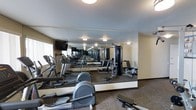 This fitness center is thoughtfully designed to support a full range of workout routines within a single-level layout. The space is outfitted with an array of cardio equipment, including treadmills, elliptical machines, and a rowing machine, all arranged to allow comfortable circulation between stations. Strength training is also well represented, with a multi-function cable machine and supporting equipment positioned toward the center and rear of the room.

A defining architectural feature is the large floor-to-ceiling mirror panel spanning a full wall, which effectively doubles the perceived depth of the room and allows users to monitor their form during exercise. The mirrored wall also reflects the lighting from the flush-mount ceiling fixtures, contributing to a bright and evenly lit environment. Neutral-toned walls and carpet flooring add a clean, functional aesthetic that keeps the focus on the equipment. A wall-mounted television is positioned for visibility across the cardio area, offering entertainment during workouts. Natural light from a window along one side supplements the overhead lighting, enhancing the overall ambiance of the space.
