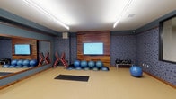 This single-floor fitness studio is thoughtfully designed to support a wide range of workout activities. The exercise area features a clean, light-toned resilient floor that provides a functional and comfortable surface for both standing exercises and floor-based routines, as evidenced by the yoga mat positioned at the center of the space.

The room is defined by bold, dark blue patterned accent walls that give the space an energetic and modern atmosphere. A standout feature is the warm wood-paneled focal wall, which serves as a backdrop for a large flat-screen display — ideal for guided workouts or fitness programming. Overhead, linear fluorescent lighting ensures the space is well-lit throughout.

Equipment is neatly organized along the perimeter of the room. A rack of red medicine balls and additional free weights are accessible along one wall, while a tiered dumbbell rack provides a range of resistance options on the opposite side. A collection of stability balls and BOSU balance trainers are lined up beneath the display screen, offering versatile options for balance and core training. A large wall mirror enhances the sense of openness and allows users to monitor their form during exercise.

Overall, this fitness studio combines practical functionality with a polished, modern aesthetic, making it a well-rounded space for diverse training needs.