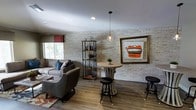 This single-floor space showcases a thoughtfully curated industrial-chic aesthetic that blends comfort with style. The interior is anchored by striking whitewashed brick accent walls, which lend a warm yet contemporary character to the room. Rich hardwood-style flooring runs throughout, providing a cohesive and polished foundation for the space.

The lounge area features a generously sized sectional sofa paired with an accent armchair, both upholstered in neutral grey tones and accented with colorful throw pillows in blue and patterned fabrics. A woven area rug grounds the seating arrangement, adding texture and warmth. A low-profile coffee table completes the conversational grouping.

Complementing the lounge seating is a bistro-style round table with a sculptural pedestal base, accompanied by industrial-design bar stools — ideal for casual conversation or individual use. An open metal-and-wood shelving unit adds both storage and decorative display space.

Overhead, a sleek pendant light with an Edison-style bulb hangs as a focal point, enhancing the industrial ambiance alongside recessed ceiling lighting. Natural light filters in through a window dressed with a Roman shade. A large, ornately framed piece of wall art adds a bold visual statement, rounding out the space's eclectic and inviting character.