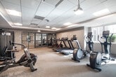 The fitness center at 2930 Rockaway Ave in Oceanside, NY is a well-appointed, single-floor facility designed to support a comprehensive workout experience. The space features a clean, modern aesthetic with neutral-toned walls, a drop-panel ceiling with recessed and pendant lighting, and a combination of carpet and hardwood-style flooring throughout.

The main exercise area is generously sized and equipped with multiple treadmills, elliptical machines, stationary bikes, and an air rowing machine, providing ample cardio options. A dedicated rack of dumbbells lines one wall, offering a range of free weights for strength training. A multi-functional cable and weight machine, along with a flat workout bench, rounds out the strength training offerings.

A separate studio room is accessible through floor-to-ceiling glass-paneled sliding doors, providing a distinct space for stretching, yoga, or group fitness activities. This room features hardwood-style flooring and textured accent walls, creating a distinct and focused workout environment.

Natural light filters in through windows along one wall, enhancing the bright and energizing atmosphere of the space. A water fountain is conveniently located near the entrance, and the facility is accessible via double glass doors. Overall, the fitness center offers a clean, functional, and inviting environment suited for a variety of exercise routines.