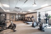 The fitness center at 2930 Rockaway Ave in Oceanside, NY is a well-appointed, single-floor facility designed to support a comprehensive workout experience. The space features a clean, modern aesthetic with neutral-toned walls, a drop-panel ceiling with recessed and pendant lighting, and a combination of carpet and hardwood-style flooring throughout.

The main exercise area is generously sized and equipped with multiple treadmills, elliptical machines, stationary bikes, and an air rowing machine, providing ample cardio options. A dedicated rack of dumbbells lines one wall, offering a range of free weights for strength training. A multi-functional cable and weight machine, along with a flat workout bench, rounds out the strength training offerings.

A separate studio room is accessible through floor-to-ceiling glass-paneled sliding doors, providing a distinct space for stretching, yoga, or group fitness activities. This room features hardwood-style flooring and textured accent walls, creating a distinct and focused workout environment.

Natural light filters in through windows along one wall, enhancing the bright and energizing atmosphere of the space. A water fountain is conveniently located near the entrance, and the facility is accessible via double glass doors. Overall, the fitness center offers a clean, functional, and inviting environment suited for a variety of exercise routines.