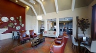 Located at 11289 E Hamilton Crest Blvd in Fishers, Indiana, this sophisticated single-floor space makes a striking impression through its bold architectural character and thoughtfully curated interiors. The central lounge area commands attention with soaring vaulted ceilings accented by crisp white exposed beams, creating an airy and grand atmosphere. A statement chandelier hangs above a comfortable seating arrangement featuring rich burgundy leather armchairs with nail-head trim, a cream upholstered sofa, and a sleek dark coffee table set atop a vibrant patterned area rug over hardwood-style flooring. Warm recessed lighting and pendant fixtures complement the natural tones throughout the space. A bold accent wall in deep red adds a dramatic focal point, while decorative wall art and metallic accents contribute to the refined aesthetic. Open sightlines connect the lounge to a well-appointed kitchen area, outfitted with dark cabinetry, mosaic tile backsplash, stainless steel appliances, and bar-height seating — ideal for entertaining or casual gatherings. Structural columns frame the transition between spaces, reinforcing the architectural cohesion. Overall, the space exudes a warm, upscale ambiance suited for community engagement and relaxation.
