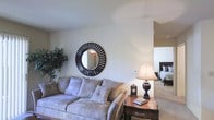 This single-floor residence offers a thoughtfully arranged layout with a warm and inviting atmosphere throughout. The living area serves as the heart of the space, showcasing soft, neutral wall tones that create a calm and relaxing environment. A plush, light gray sofa anchors the seating area and is complemented by an assortment of coordinating cushions for added comfort. A decorative round mirror with a bold sunburst frame adds a striking focal point to the main wall, while a lush indoor plant in the corner introduces a refreshing touch of greenery. A richly stained wooden side table sits adjacent to the sofa, styled with a classic table lamp, a small decorative clock, and a decorative urn, lending the space an air of traditional elegance. The open layout allows for a natural visual flow into the bedroom, which is partially visible in the background and features consistent neutral carpeting that ties the spaces together cohesively. The overall ambiance is one of understated sophistication — clean, uncluttered, and designed for comfortable everyday living. Soft, even lighting throughout enhances the welcoming character of the space.