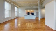 This single-floor urban loft showcases a compelling blend of industrial and classic architectural elements. The expansive open-plan living area immediately impresses with soaring ceilings, exposed ductwork and beams, and a row of large windows that flood the space with natural light. Elegant decorative columns with ornate capitals anchor the room, adding a distinctive character. Rich, warm hardwood floors run throughout the main living areas, further enhancing the inviting atmosphere. A white-mantled fireplace serves as a focal point in the living room, complemented by views of the surrounding cityscape through generous windows.

The kitchen is thoughtfully designed as a central island unit, featuring warm wood cabinetry, dark countertops, a tile backsplash, and black appliances including a range, microwave, and refrigerator. Pendant lighting above the island adds a refined touch.

The bedroom is generously proportioned, carpeted for comfort, and distinguished by a striking arched window that frames outdoor views and admits abundant natural light. The room's elevated ceilings, visible ductwork, and recessed lighting continue the loft's cohesive industrial-chic aesthetic.

The bathroom is well-appointed with a distinctive angled glass-enclosed corner shower, tile flooring, a wood-toned vanity with a single sink, and a large mirror, presenting a clean and functional design.