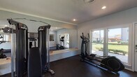 This single-level fitness center is thoughtfully designed to support a comprehensive workout experience. The space is finished with durable rubber flooring throughout, providing both safety and comfort during exercise. Recessed lighting illuminates the room evenly, complemented by natural light streaming through multiple windows and a glass door that offers views of the surrounding outdoor greenery.

The workout area is equipped with a varied selection of exercise equipment, including a treadmill, two elliptical machines, and an Inspire-brand cable machine suitable for a wide range of strength training exercises. A multi-tier dumbbell rack holds an organized selection of free weights, catering to various fitness levels. A wall-mounted flat-screen television adds entertainment options during workouts.

Large wall mirrors span a significant portion of one wall, visually expanding the space and allowing users to monitor their form. A white brick accent detail along the base of the mirrored wall adds a subtle architectural touch. The neutral white walls and clean lines contribute to an open, uncluttered atmosphere.

An adjacent passthrough window and glass-panel door connect the fitness area to what appears to be a leasing or common area, suggesting this facility is part of a larger community property.