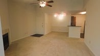 This single-floor space offers a clean, open-concept layout that seamlessly connects the main living area to the kitchen. The living room is generously sized, finished with light-toned wall-to-wall carpeting that lends a warm, neutral feel throughout. A ceiling fan with an integrated light fixture is centrally mounted, providing both air circulation and ambient illumination. Natural light enters through the space, complementing the track lighting installed near the kitchen transition area.

The entry features a classic paneled white door with a dark floor mat, offering a welcoming point of arrival. An open passage leads directly into the main living area, which provides ample room for a variety of furniture arrangements.

Adjacent to the living area, the kitchen is defined by a raised breakfast bar countertop in a dark finish, creating a natural visual divider while maintaining the open flow of the space. The kitchen area includes cabinetry and appears to feature additional appliances, making it both functional and accessible. The light-colored walls throughout contribute to a bright, airy ambiance, and the overall layout is practical and versatile, well-suited for everyday living.