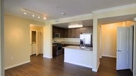 This single-story space showcases an open-concept layout that seamlessly connects the kitchen and living areas. The kitchen is a standout feature, equipped with stainless steel appliances including a gas range, built-in microwave, and a French door refrigerator. Rich, dark wood cabinetry lines the walls and extends to the ceiling, complemented by granite countertops that add a touch of elegance. A peninsula with a built-in sink and matching granite surface provides additional workspace and casual seating potential, while also serving as a natural divider between the kitchen and the adjoining living area. Crown molding along the ceiling adds architectural refinement throughout, and track lighting in the kitchen area ensures ample task illumination. The warm-toned hardwood flooring flows continuously through the open space, enhancing the cohesive aesthetic. Visible from the main area, a dedicated laundry area with a washer and dryer offers added convenience. The light-colored walls and neutral palette throughout contribute to a bright and welcoming atmosphere.