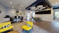 Located at 201 Crater Circle in Petersburg, Virginia, this single-story community clubhouse is designed with a contemporary aesthetic that balances functionality and style. The building's interior is characterized by a cohesive color palette of navy, teal, gray, and yellow accents, creating a vibrant yet professional atmosphere throughout.

The main hall features soaring vaulted ceilings with clerestory windows that flood the space with natural light, complemented by wide-plank hardwood-style flooring. A welcoming entry foyer offers seating with bold yellow accent chairs arranged around a circular table. The central open-plan area serves as a business center, equipped with computer workstations, pendant lighting, and an ornate laser-cut room divider that defines a private consultation nook with a wall-mounted display and conference seating.

An adjoining multipurpose lounge and dining area features a striking navy ceiling with geometric hexagonal acoustic panels, pendant lighting, dual wall-mounted televisions, a live-edge wood communal table with teal chairs, and a built-in beverage and refreshment station along the wall.

A separate lounge area provides comfortable seating with a brown sofa and plaid accent chairs accented by teal throw pillows, bathed in natural light from large bay windows overlooking the surrounding community.

The fully equipped fitness center, with bold teal walls and striped carpet flooring, houses cardio machines, free weights, a cable machine, weight benches, and mirrored walls, with ample natural light provided by multiple windows.