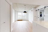 Located at 2198 Boulevard De Maisonneuve Est in Montréal, this thoughtfully designed one-bedroom, one-bathroom unit blends industrial character with contemporary finishes. The open-concept living area is anchored by striking exposed concrete ceilings and light hardwood flooring throughout, creating a warm contrast to the crisp white walls. A large window floods the living space with natural light and frames views of the surrounding Montréal cityscape.

The kitchen is a functional centerpiece, featuring a sleek white waterfall island with an integrated sink, white cabinetry, subway tile backsplash, stainless steel appliances including a full-size refrigerator and built-in microwave, and track lighting overhead. A stylish black-framed sliding barn door connects the main living area to the bedroom, adding an architectural detail that enhances the modern aesthetic.

The bedroom is a bright, well-proportioned retreat with continued hardwood flooring, a built-in closet, and a large window overlooking the city. The all-white bathroom is efficiently laid out with a floating vanity, glass-enclosed shower, and a stacked washer-dryer unit conveniently integrated into the space. This unit offers a clean, urban living experience in the heart of Montréal.