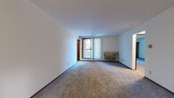 This single-floor unit is located on Milwaukee's East Side at 839–899 E State St, offering a practical and well-organized layout across several distinct spaces. The living area is generously proportioned, featuring neutral wall-to-wall carpeting, white walls, and a large window dressed with vertical blinds that allows natural light to fill the room. A wall-mounted HVAC unit provides climate control, and an entry door with warm wood tones anchors one end of the space.

The kitchen is equipped with medium-toned wood cabinetry offering substantial storage, dark granite-style countertops, a double stainless steel sink, and black appliances including a dishwasher and range. Light tile flooring contrasts with the cabinetry for a clean, functional aesthetic.

The bedroom features carpeted flooring, a large sliding wood-panel closet, and a window with views of surrounding urban greenery and buildings. An additional carpeted room with a window and wall-unit AC offers flexible use as a second bedroom or office.

The bathroom includes a vanity with wood cabinetry, a large mirror with overhead lighting, tile flooring, and a walk-in shower with sliding glass doors and tiled surround. A dedicated laundry area with a stacked washer and dryer adds notable convenience to the overall layout.