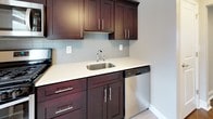 This single-floor space combines classic character with modern updates throughout its well-appointed rooms. Rich, dark hardwood floors run consistently across the living and bedroom areas, reflecting light and lending warmth to the neutral gray and beige-toned walls. The living area is generously proportioned, featuring a large window fitted with blinds, a baseboard radiator, and a paneled entry door with sidelights that allows natural light to filter in.

The bedrooms are spacious and versatile, each featuring the same polished hardwood flooring. One bedroom includes a closet with built-in hanging rod behind a classic paneled door, while another is lit by recessed ceiling fixtures and connects to adjacent rooms through a bright doorway. Additional windows in the bedrooms invite outdoor views and ample daylight.

The kitchen is thoughtfully outfitted with rich, dark mahogany-toned cabinetry, light quartz countertops, a stainless steel sink, and a glass tile backsplash. A stainless steel dishwasher adds modern convenience.

The bathroom features a clean, tiled design with white subway tiles accented by dark border tiles, a soaking tub, a window for natural ventilation, and a gray slate-style floor — creating a sleek, functional space.