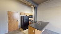 Located at 188 N Water St in the heart of Rochester, NY, this urban loft-style unit showcases a compelling blend of industrial character and functional living. The space is defined by notably high ceilings that amplify the sense of openness throughout, while exposed structural columns, visible ductwork, and track lighting overhead reinforce the building's converted industrial heritage.

The kitchen is efficiently designed with light wood cabinetry, a stainless steel gas range, a black refrigerator, and a built-in microwave. A peninsula with a dark countertop and integrated sink serves as a natural divider, transitioning seamlessly into the open living area. The living space features wall-to-wall carpet and benefits from natural light streaming through large, multi-pane windows that frame views of the surrounding streetscape and greenery.

The bedroom is a well-proportioned, carpeted room with its own multi-pane window offering outdoor views and baseboard heating along the perimeter wall, ensuring year-round comfort.

The bathroom is finished with light ceramic tile flooring, a warm wood vanity with a dark countertop, a wall-mounted mirror with built-in lighting, and a full bathtub and shower combination. Light wood doors are consistent throughout the unit, contributing to a cohesive, modern aesthetic. The overall atmosphere is urban, open, and rich with architectural detail.