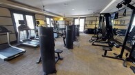 This fitness center offers a thoughtfully arranged and fully equipped workout facility designed to support a wide range of exercise routines. The space is outfitted with a lineup of Paramount strength-training machines, providing dedicated stations for targeted muscle group exercises. A row of cardio equipment, including treadmills and elliptical machines, lines the perimeter near the windows, allowing users to exercise with the benefit of natural light. A multi-station cable and weight machine anchors one side of the room, ideal for versatile strength training. The flooring consists of textured carpet tile, providing a practical and durable surface suited to a fitness environment. Ceiling fans are mounted overhead, contributing to air circulation throughout the space and enhancing comfort during workouts. The walls feature a warm yellow and neutral tone color scheme, giving the room an energetic yet welcoming ambiance. Windows along multiple walls invite natural light deep into the interior, while a glass door at the rear provides additional brightness and a connection to the outdoors. The overall layout is open and organized, allowing for easy movement between equipment stations.