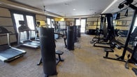 This fitness center offers a thoughtfully arranged and fully equipped workout facility designed to support a wide range of exercise routines. The space is outfitted with a lineup of Paramount strength-training machines, providing dedicated stations for targeted muscle group exercises. A row of cardio equipment, including treadmills and elliptical machines, lines the perimeter near the windows, allowing users to exercise with the benefit of natural light. A multi-station cable and weight machine anchors one side of the room, ideal for versatile strength training. The flooring consists of textured carpet tile, providing a practical and durable surface suited to a fitness environment. Ceiling fans are mounted overhead, contributing to air circulation throughout the space and enhancing comfort during workouts. The walls feature a warm yellow and neutral tone color scheme, giving the room an energetic yet welcoming ambiance. Windows along multiple walls invite natural light deep into the interior, while a glass door at the rear provides additional brightness and a connection to the outdoors. The overall layout is open and organized, allowing for easy movement between equipment stations.