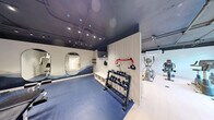 Located at 2645 Battleford Rd in Mississauga, Ontario, this contemporary fitness facility is thoughtfully designed to deliver a complete workout experience across a single floor. The main exercise area features light hardwood-style flooring and an exposed dark ceiling with industrial-style track lighting, lending the space a sleek, modern aesthetic. A subtly patterned botanical wall mural serves as a striking backdrop, while large windows along one wall invite natural light and offer views of surrounding greenery, creating an energizing atmosphere. The cardio zone is equipped with treadmills, an elliptical trainer, and stationary bikes, all arranged to maximize space and airflow. A dedicated strength and free weights area transitions to blue rubberized flooring for safety and comfort, and features a full dumbbell rack, wall-mounted resistance accessories, and built-in storage benching with a wall-mounted television. Oval-shaped mirrors are thoughtfully positioned throughout to enhance depth and functionality. The facility also includes a well-appointed washroom with large-format floor tiles, a stone-topped vanity with light wood cabinetry, matte black fixtures, an oval mirror, and a textured tile accent wall illuminated by contemporary sconce lighting — combining practicality with refined design.