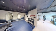 Located at 2645 Battleford Rd in Mississauga, Ontario, this contemporary fitness facility is thoughtfully designed to deliver a complete workout experience across a single floor. The main exercise area features light hardwood-style flooring and an exposed dark ceiling with industrial-style track lighting, lending the space a sleek, modern aesthetic. A subtly patterned botanical wall mural serves as a striking backdrop, while large windows along one wall invite natural light and offer views of surrounding greenery, creating an energizing atmosphere. The cardio zone is equipped with treadmills, an elliptical trainer, and stationary bikes, all arranged to maximize space and airflow. A dedicated strength and free weights area transitions to blue rubberized flooring for safety and comfort, and features a full dumbbell rack, wall-mounted resistance accessories, and built-in storage benching with a wall-mounted television. Oval-shaped mirrors are thoughtfully positioned throughout to enhance depth and functionality. The facility also includes a well-appointed washroom with large-format floor tiles, a stone-topped vanity with light wood cabinetry, matte black fixtures, an oval mirror, and a textured tile accent wall illuminated by contemporary sconce lighting — combining practicality with refined design.