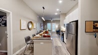 This well-appointed single-floor unit offers five bedrooms and four bathrooms designed with a clean, contemporary aesthetic. The open-concept kitchen and living area serves as the heart of the home, featuring tall ceilings that enhance the sense of space. The kitchen is outfitted with stainless steel appliances—including a refrigerator, range, and microwave—complemented by sleek white cabinetry, dark countertops, and a large island with seating, illuminated by modern pendant lighting. Light wood-tone flooring runs throughout the main living areas, adding warmth and cohesion. The living room benefits from multiple windows and a glass door that invite natural light and provide outdoor views, creating a bright and welcoming atmosphere. Each bedroom is carpeted for comfort, with windows that allow natural light in; bedrooms include closets with built-in shelving for ample storage. The four bathrooms are finished in neutral tones with tiled tub-shower combinations, modern vanities, and wood-look flooring. Neutral warm-gray walls throughout the unit create a cohesive, calming palette. Recessed lighting and ceiling fans add both functionality and style, making this a practical and modern living space suited for shared or family occupancy.