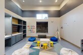 Located at 14698 Briar Forest Drive in Houston, Texas, this dedicated children's playroom is a thoughtfully designed, single-level space that offers a stimulating and engaging environment for young children. The room features soaring ceilings with a bold two-tone color scheme — dark charcoal gray on the upper walls and crisp white below — creating a striking visual contrast that defines the space's modern aesthetic. Recessed lighting illuminates the interior, while a high clerestory window allows natural light to filter in.

At the heart of the room, a colorful geometric-patterned area rug anchors a central play zone furnished with a child-sized white table, bright yellow stools, and an assortment of bean bag seats in blue and white. A canvas teepee sits in one corner, providing a cozy nook for imaginative play, while cheerful nature-themed wall decals and alphabet lettering add educational and decorative character to the walls.

A full wall of built-in gray and yellow shelving with lower cabinetry and an integrated television provides ample storage and entertainment options. Additional toy organizers and play equipment complete the setup. A bold "PLAY" wall sign near glass-panel doors — which connect to an adjacent fitness center — reinforces the room's playful purpose and sense of community amenity.