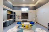 Located at 14698 Briar Forest Drive in Houston, Texas, this dedicated children's playroom is a thoughtfully designed, single-level space that offers a stimulating and engaging environment for young children. The room features soaring ceilings with a bold two-tone color scheme — dark charcoal gray on the upper walls and crisp white below — creating a striking visual contrast that defines the space's modern aesthetic. Recessed lighting illuminates the interior, while a high clerestory window allows natural light to filter in.

At the heart of the room, a colorful geometric-patterned area rug anchors a central play zone furnished with a child-sized white table, bright yellow stools, and an assortment of bean bag seats in blue and white. A canvas teepee sits in one corner, providing a cozy nook for imaginative play, while cheerful nature-themed wall decals and alphabet lettering add educational and decorative character to the walls.

A full wall of built-in gray and yellow shelving with lower cabinetry and an integrated television provides ample storage and entertainment options. Additional toy organizers and play equipment complete the setup. A bold "PLAY" wall sign near glass-panel doors — which connect to an adjacent fitness center — reinforces the room's playful purpose and sense of community amenity.