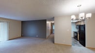 This single-floor residence offers a thoughtfully designed layout that balances open communal spaces with comfortable private rooms. The expansive living room is highlighted by plush carpeting, warm neutral tones, and a large sliding glass door that floods the space with natural light and provides views of the surrounding greenery. An accent wall in a bold charcoal-gray hue adds a contemporary touch to the living area.

The open-concept layout connects the living room to the dining area, which is illuminated by an elegant chandelier, and flows seamlessly into the kitchen. The kitchen features light wood cabinetry, granite-style countertops, tile flooring, and black appliances including a range and over-the-range microwave, along with a pass-through that maintains visual connectivity with the main living space.

The bedrooms are generously sized with soft carpeting, ceiling fans with light fixtures, and windows fitted with horizontal blinds that allow for controlled natural light. One bedroom includes a built-in window seat, adding a charming architectural detail. The primary bedroom provides direct access to a closet and a full bathroom.

The bathroom is well-appointed with a single-sink vanity, wood cabinetry, a large mirror with above-mount lighting, and a full tub-shower combination with a curved rod. Textured ceilings and warm wood baseboards are consistent throughout, lending the space a cohesive and inviting character.